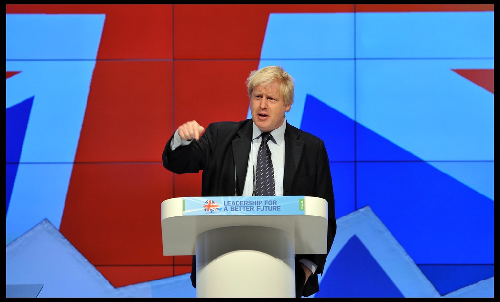 boris johnson addressing a crowd on a podium with a colourful background