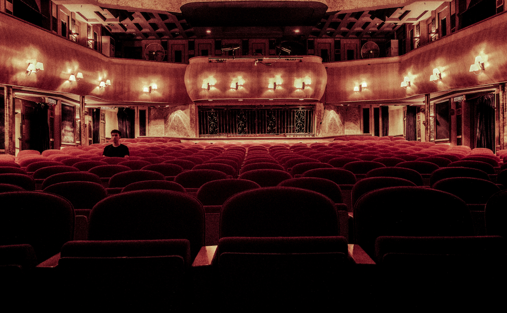 A dimly lit red theatre auditorium, with one single person sitting alone in the audience.
