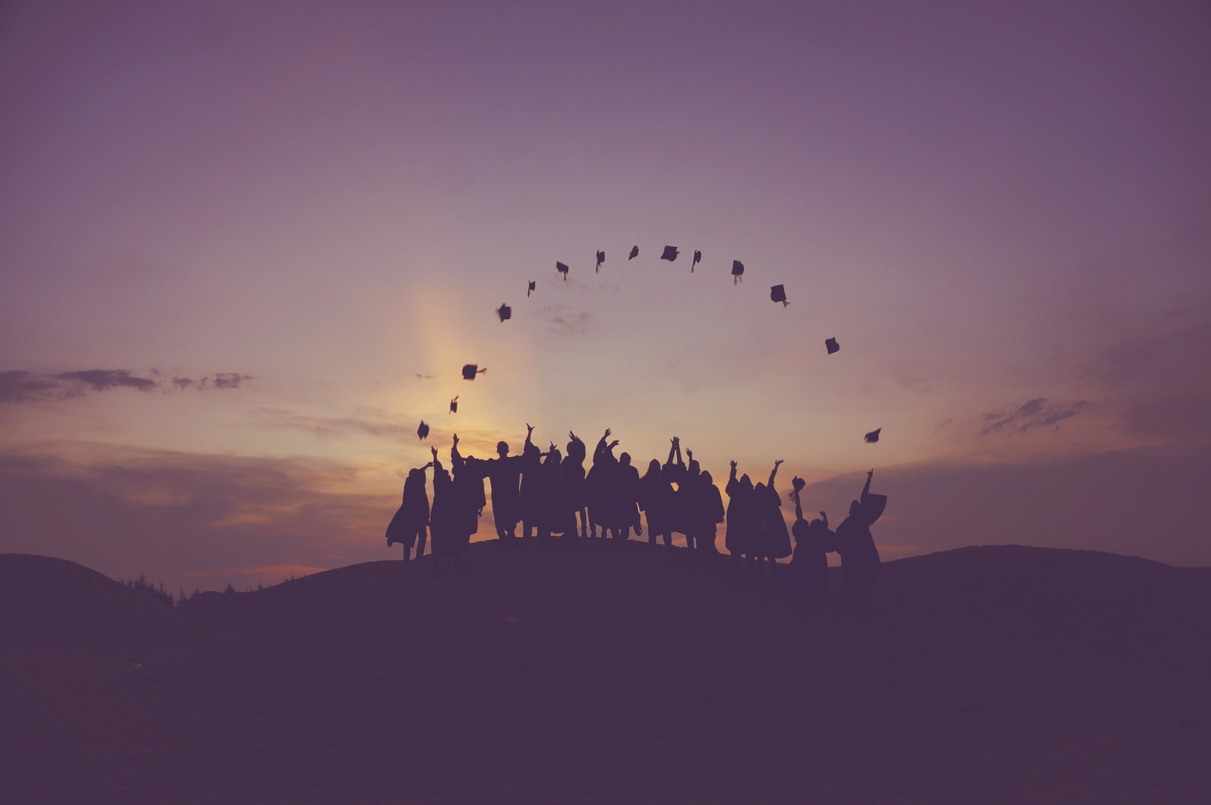 A silhouette of a group of graduates in gowns throwing their caps up in the air in front of a sunset.