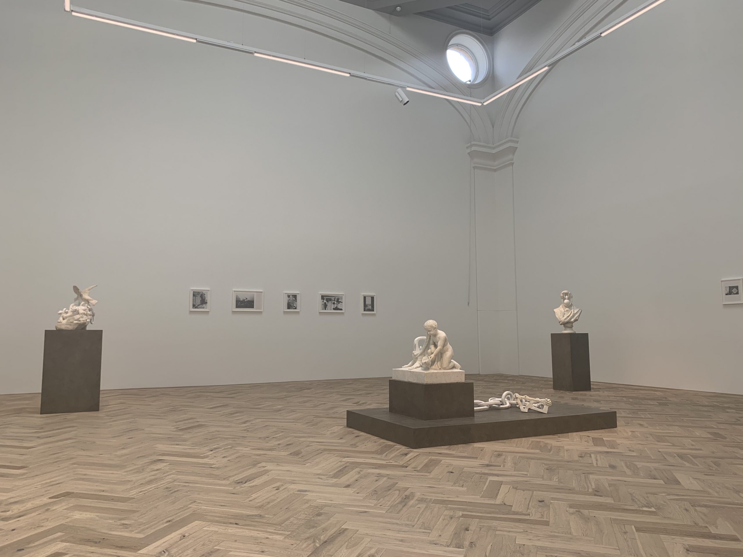 Exhibition space with white wall and wooden floor. In the middle of the room over three plinth there are three white marble sculptures. On the wall five drawings are framed