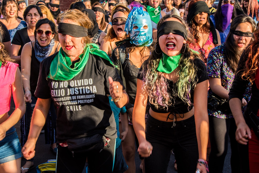 Group of people with a black blindfold, multiple with a green bandana around their neck chanting and performing 'A Rapist on Your Path'