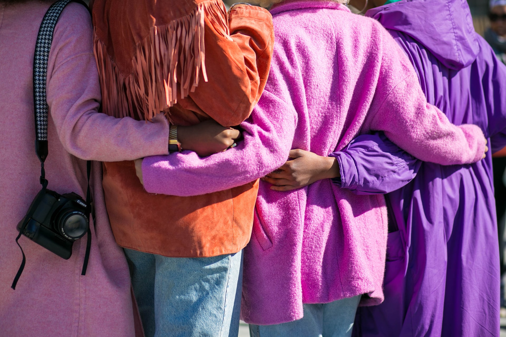 The picture is of 4 people's backs, they wear colourful coats in pink,orange, pink and purple as they have their arms around each others' backs.