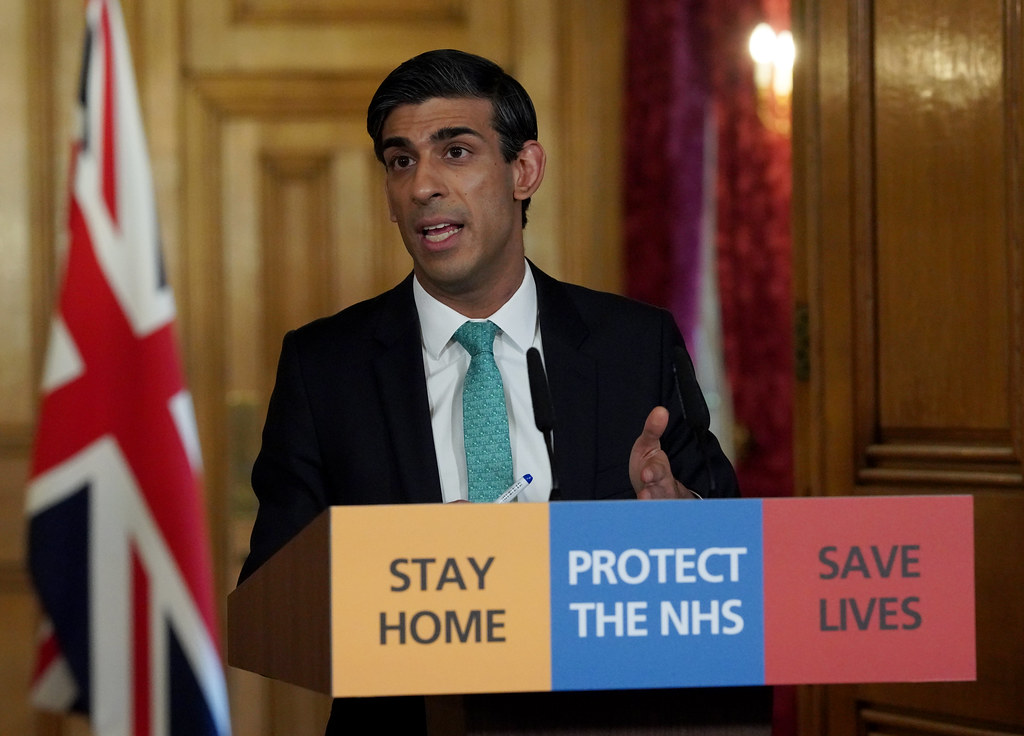 chancellor rishi sunak at a podium with a sign that says stay home protect the nhs save lives