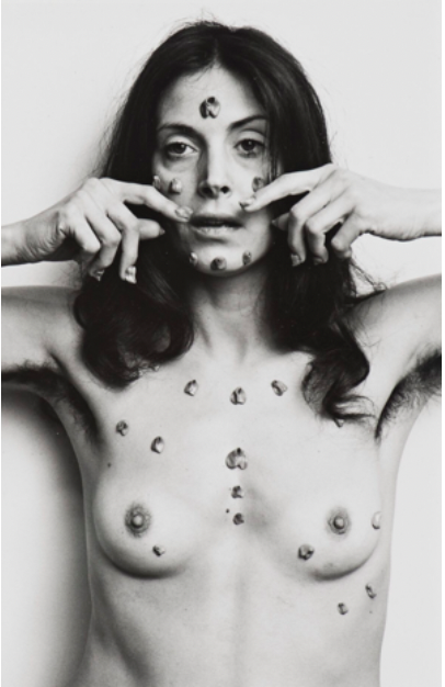 Black and white photograph of a naked woman with dark hair reaching her shoulders. On her face and chest there are little chewing gum shaped like vulvas