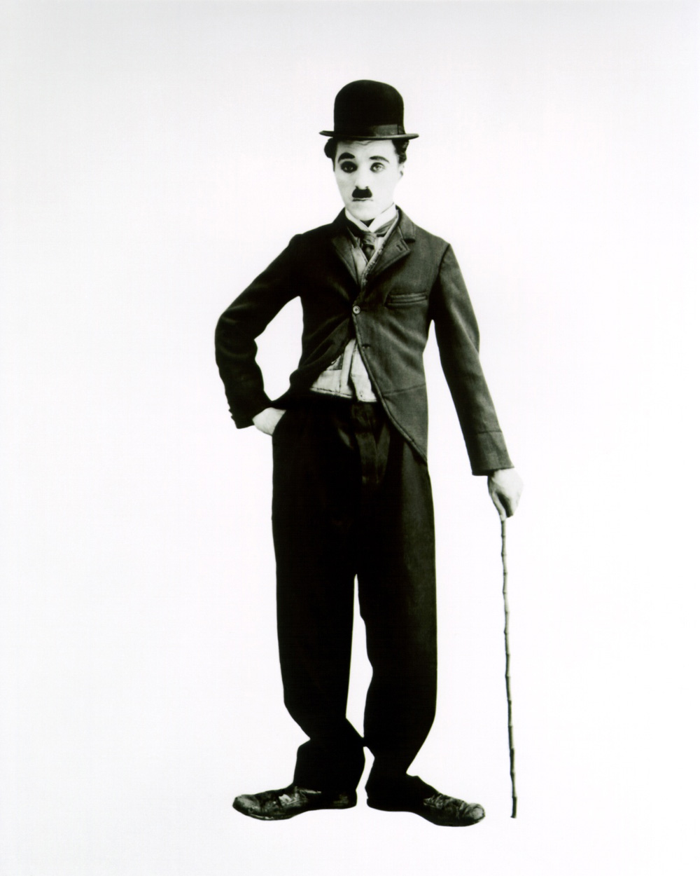 Black and white image of Charlie Chaplin standing with feet out-turned, hand on hip and holding a cane.