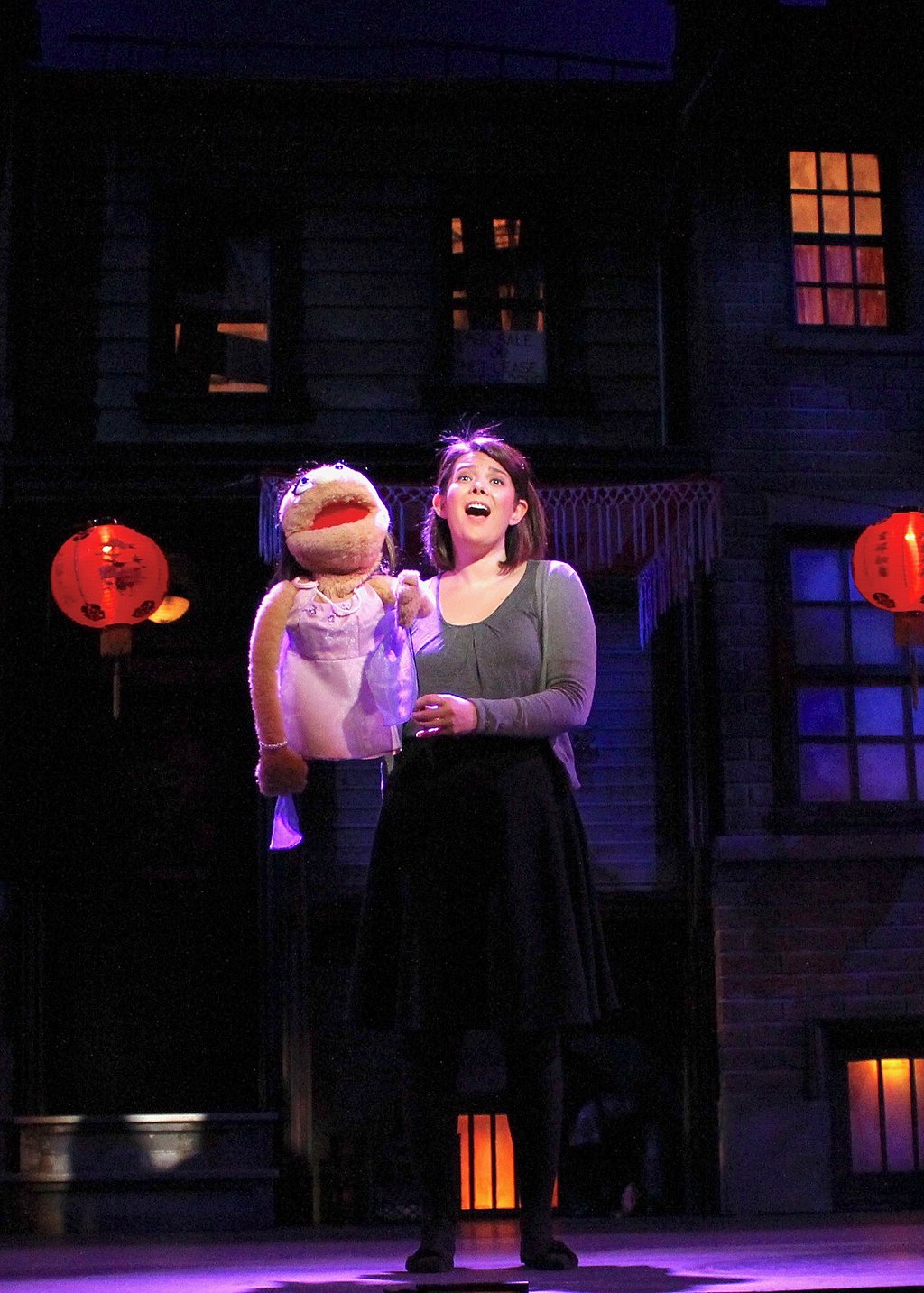 An actress performs with a puppet in front of a dark background.