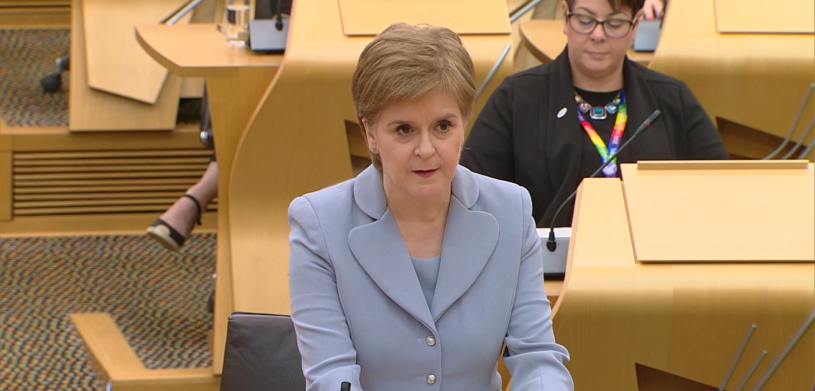 Image of first minister Nicola Sturgeon speaking at Scottish Parliament. She is standing and wearing a light blue suit