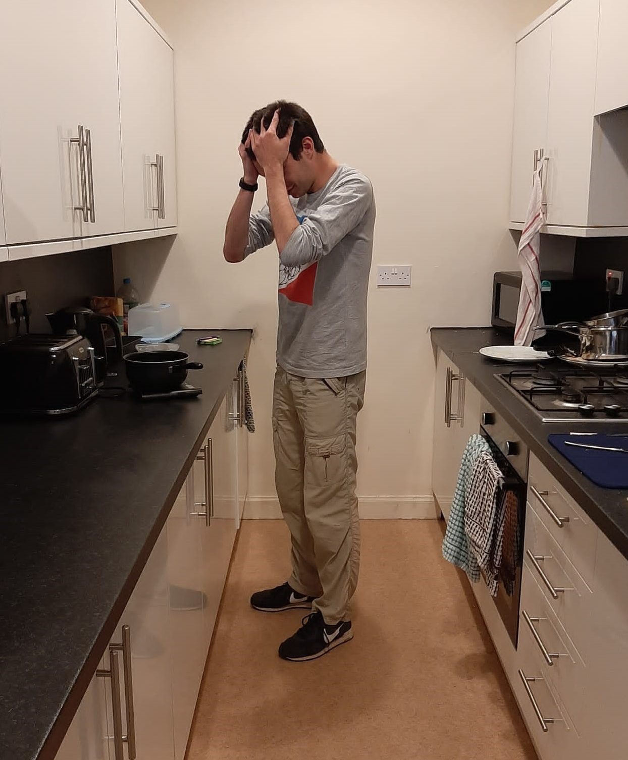 A man stands over a saucepan in a kitchen, his head in his hands.