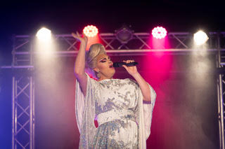 Star of Life’s A Drag on stage holding a microphone and singing. They are wearing a white dress with sleeves that drape and their hair is up in a beehive fashion.