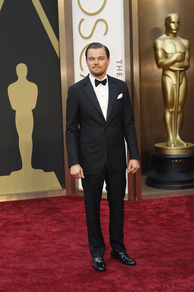 Leonardo DiCaprio stands on the red carpet at the Oscars. He is wearing a black tuxedo and staring at cameras