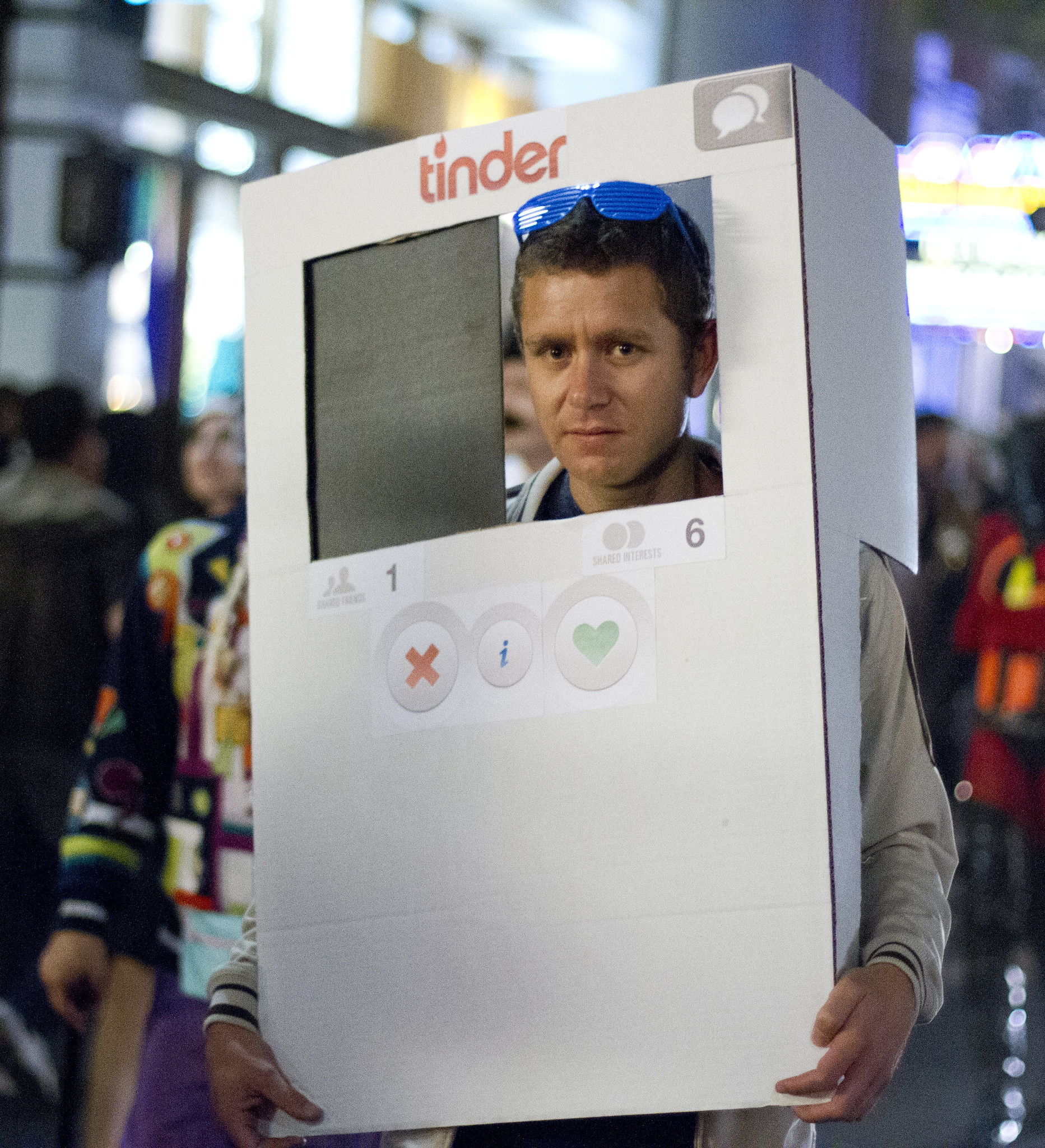 A relatively dour looking man wearing a costume resembling a phone displaying the Tinder application, with the cutout for the head being situated above the "Swipe Left" and "Swipe Right" buttons.