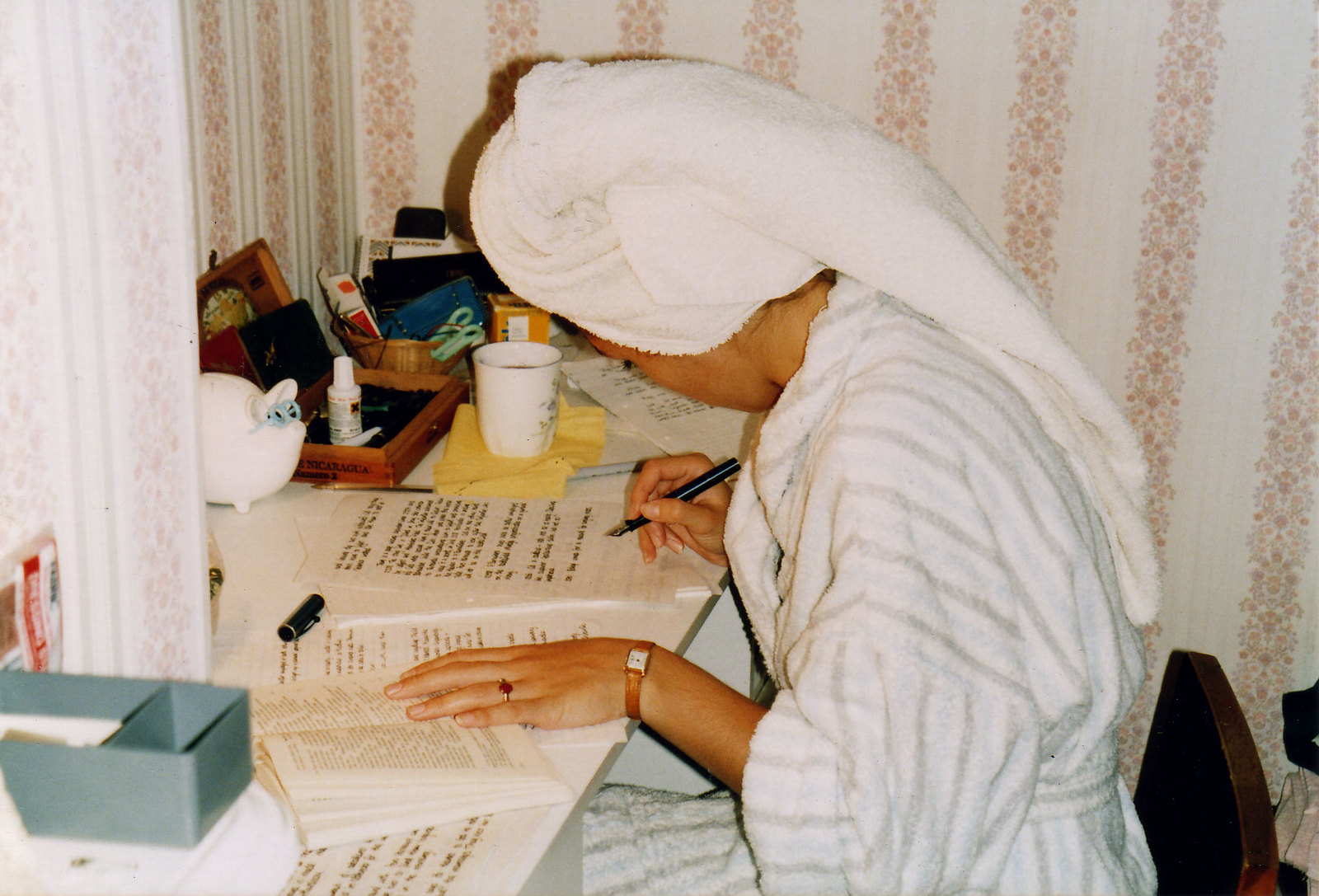 woman with towel wrapped round head and in striped dressing gown sits at her desk as she studies