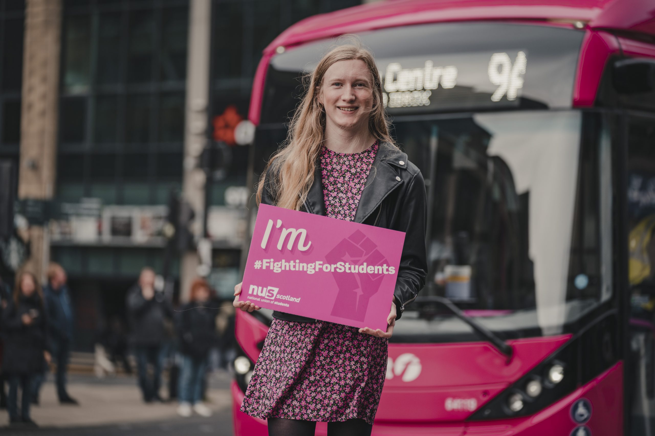 A woman, standing in front of a bus with a sign reading "I'm #FightingForStudents"
