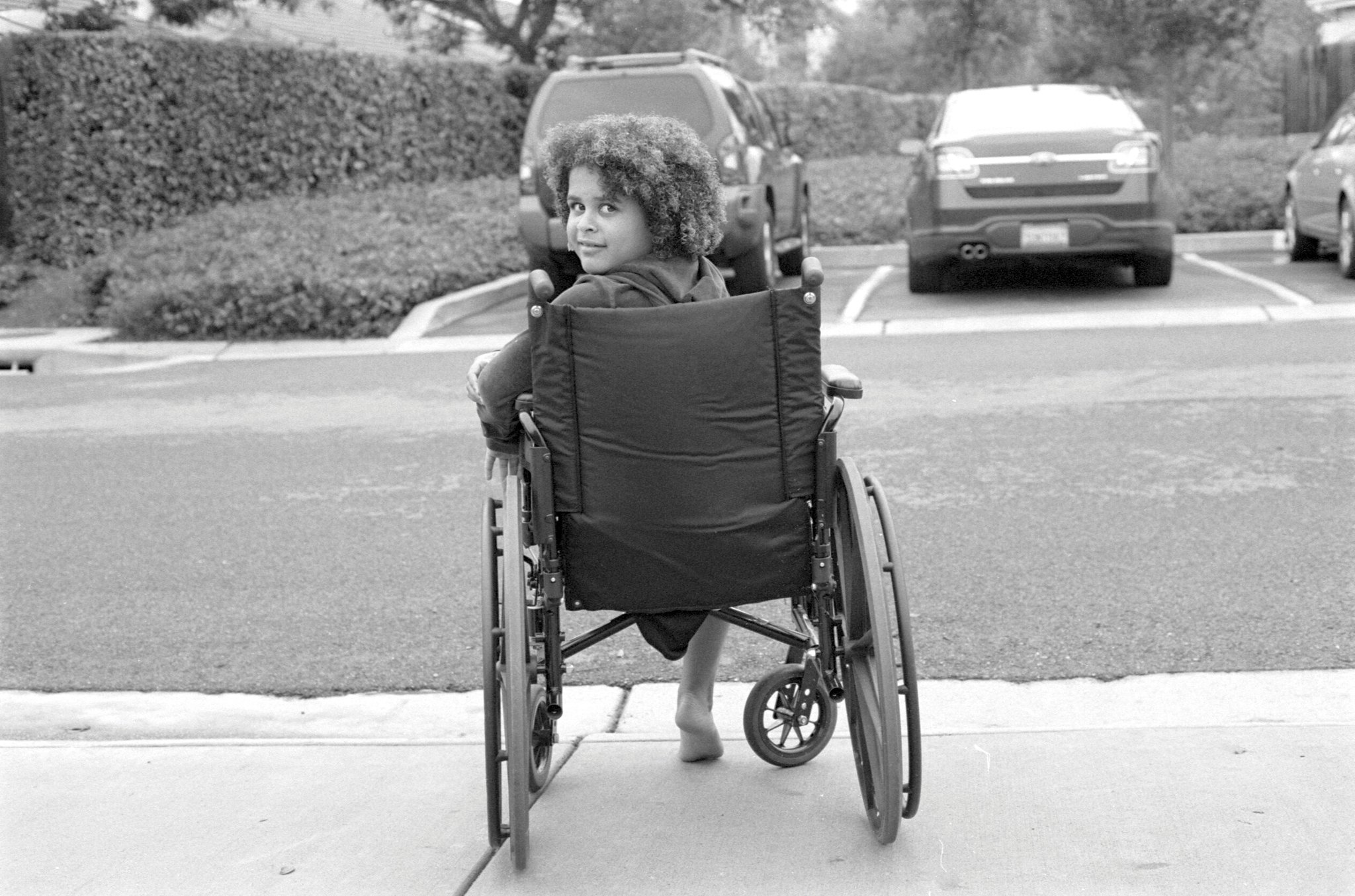 A child sits in a wheelchair, which is facing away from the camera. They are turning around and facing the camera. Image is in black and white.