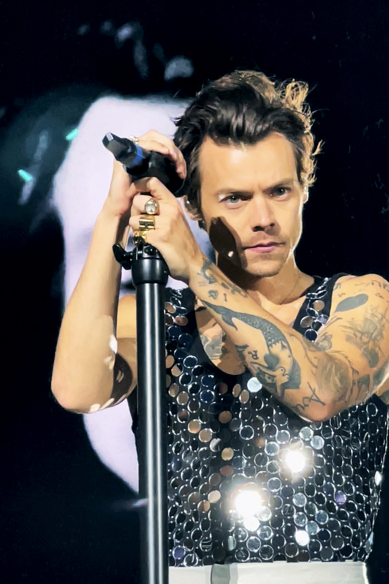 Harry Styles stood in front of a microphone on stage at Wembley. We is wearing a sequin vest top.