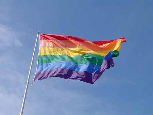 Rainbow Pride Flag flying at full mast. The sky is a clear blue.