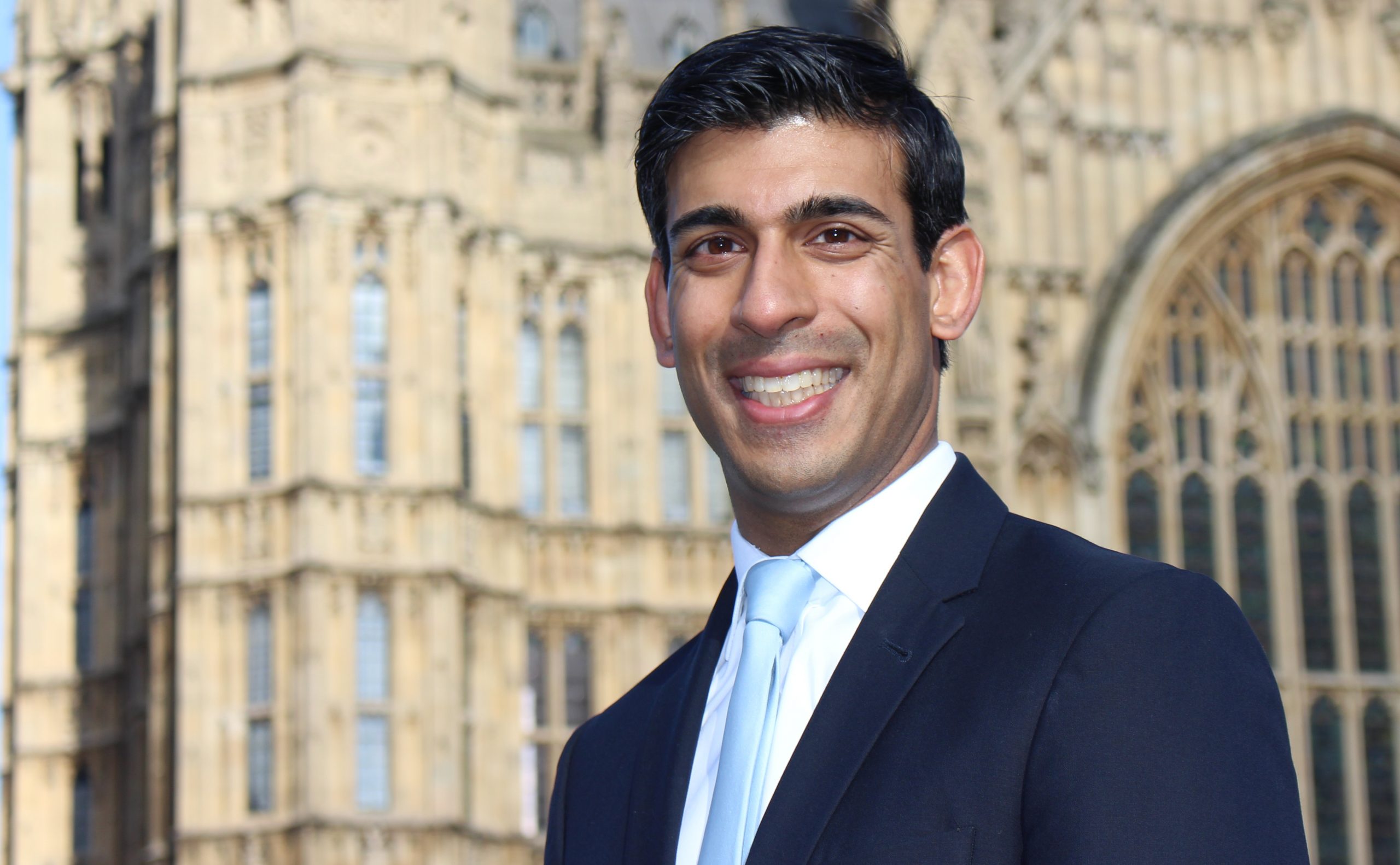 Rishi Sunak smiling at the camera. He is wearing a black suit with a light blue tie. Behind him is the Houses of Parliament.