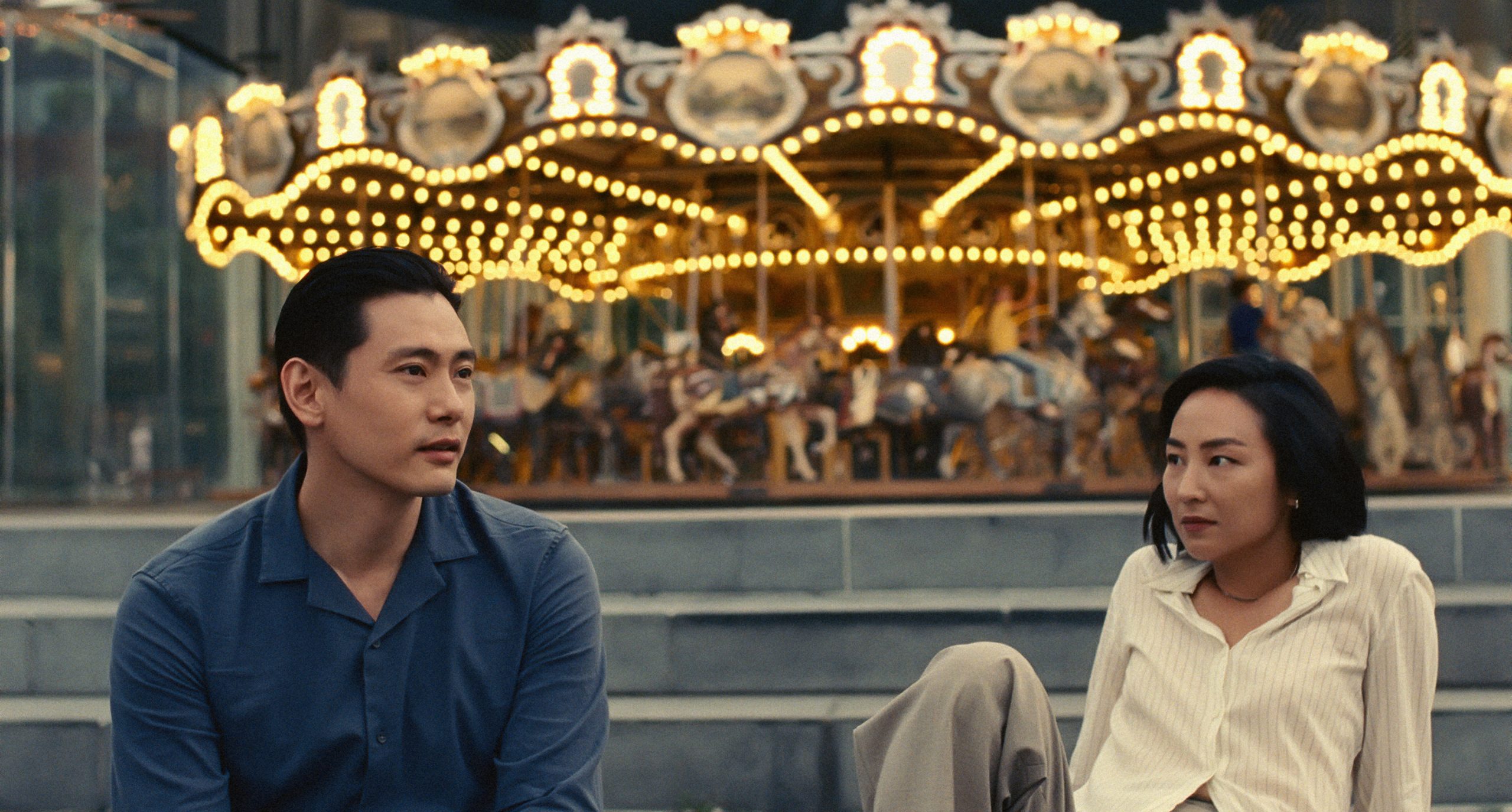 A man and a woman sit in front of a fairground carousel looking at each other.