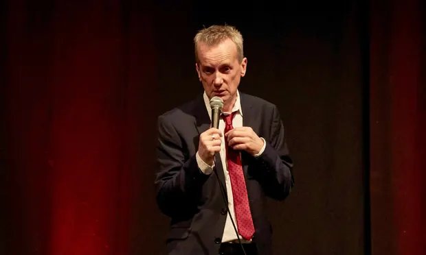 Frank Skinner stands in a suit and tie, holding a microphone