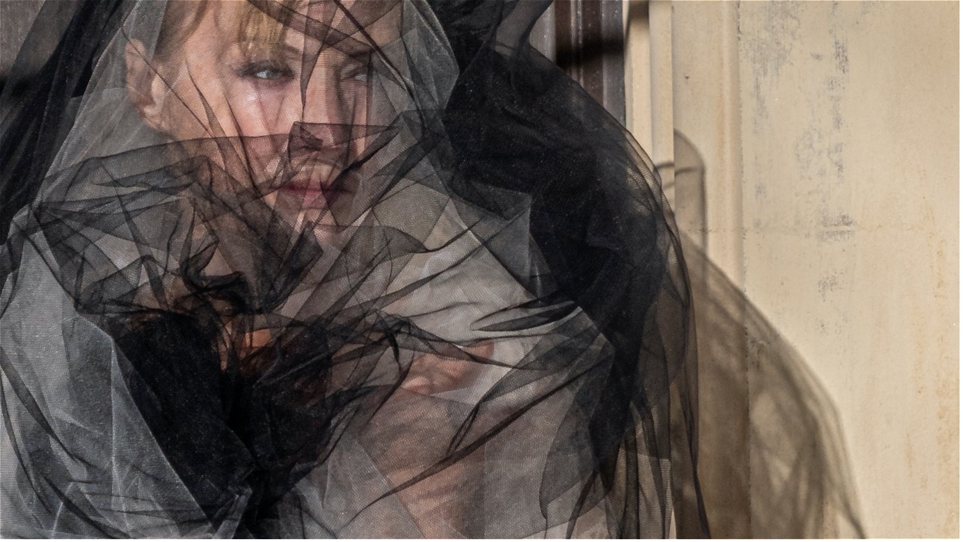 Havisham peering through a black veil, against a tan background.