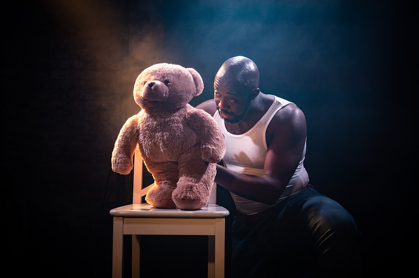 Actor crouches behind a chair with a Teddy bear on, in dramatic stage lighting.