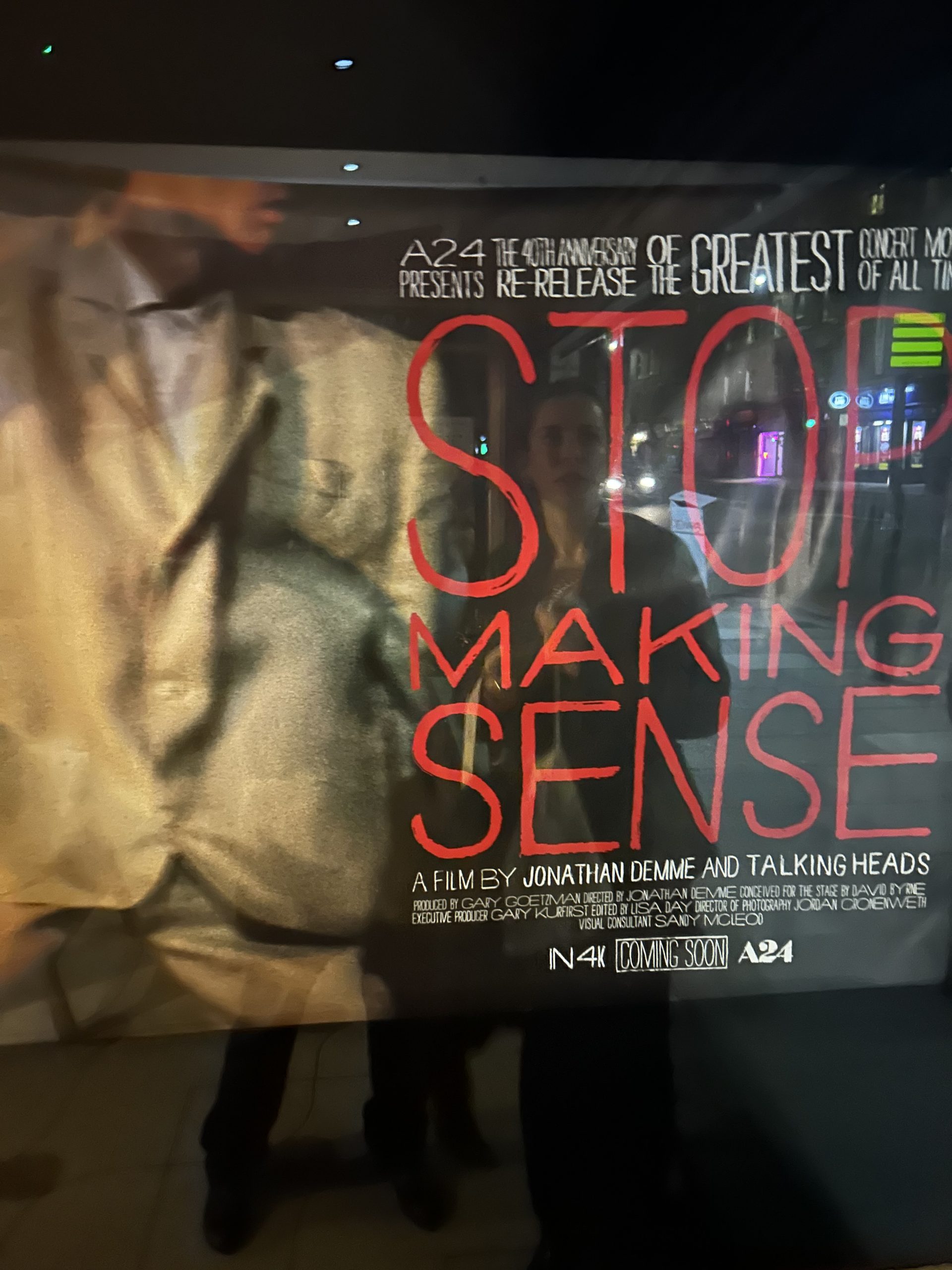 This is the image of the movie poster for 'Stop Making Sense'. We can see the white suit of David Byrnes, with red lettering. This is a picture of the poster, so we can see the reflection of the street on it.
