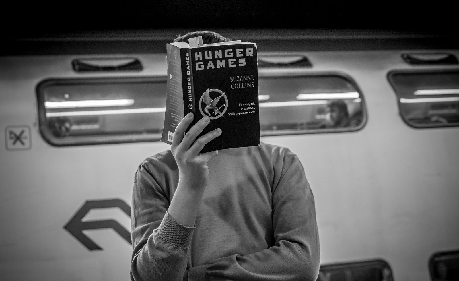 black and white picture of a man standing in front of a moving train, holding The Hunger Games book over his face