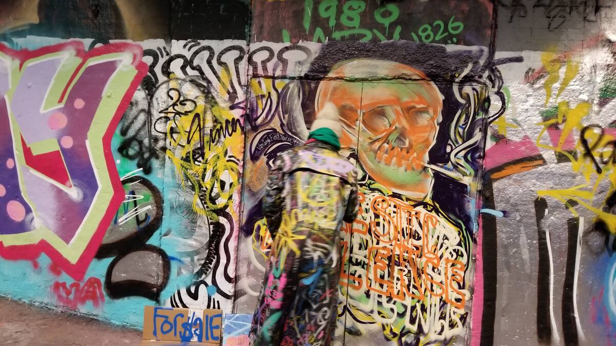 Walter DeForest painting graffiti on a wall in Edinburgh. The artwork is a skull with many colours and abstract shapes surrounding it.