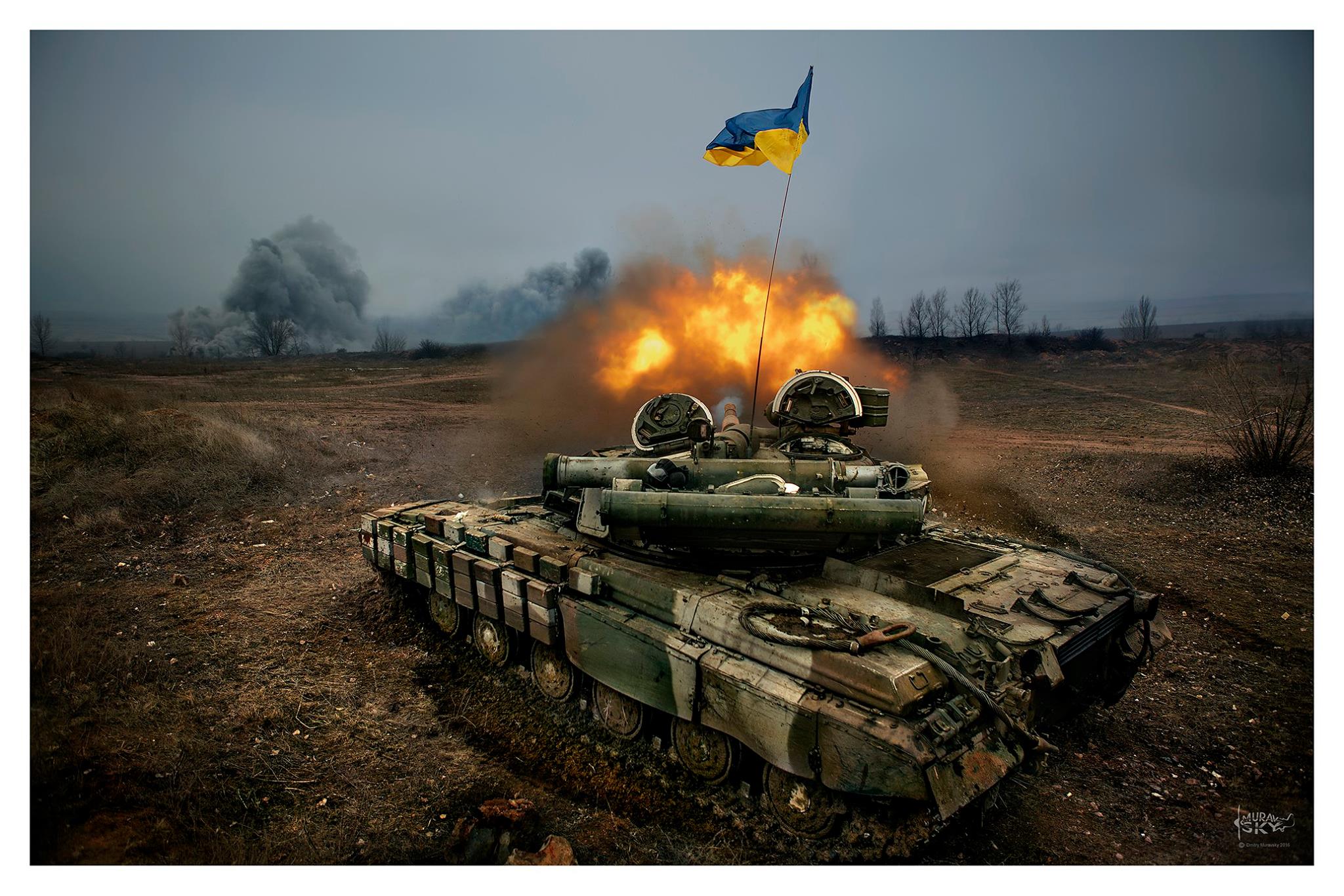 Camo-coloured tank with Ukraine flag firing a round on barren land